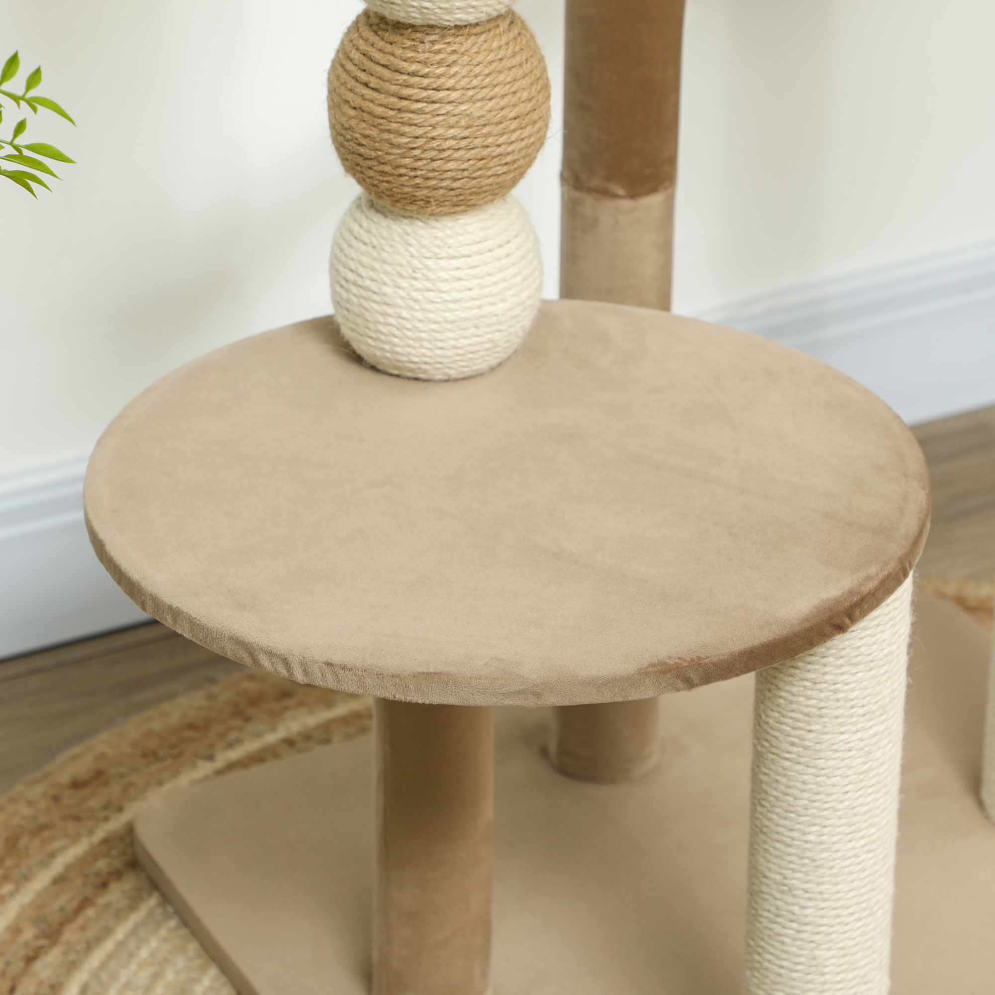 Árbol para gatos con plataforma beige, postes y bolas envueltas en cuerda. Una alfombra redonda está en la base. Hojas verdes arriba a la izquierda.