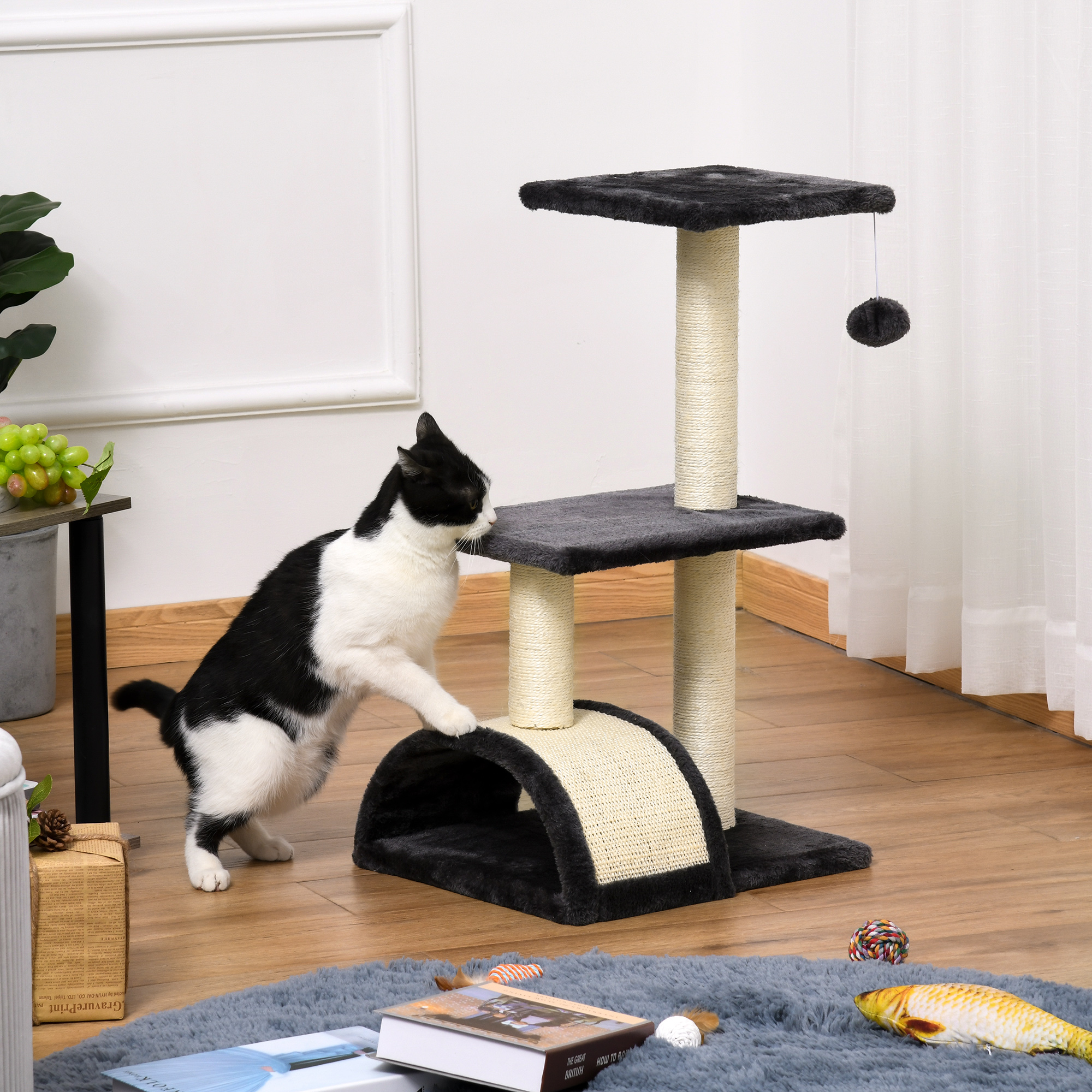 Un gato blanco y negro juega en un rascador. El árbol tiene postes para rascar, plataformas y un juguete colgante. Una alfombra gris está en el suelo.