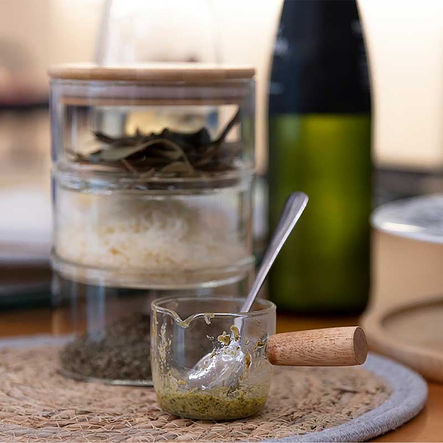 Un barattolo di vetro con spezie, un bicchierino con manico in legno e pesto, un cucchiaio e una bottiglia verde sullo sfondo.