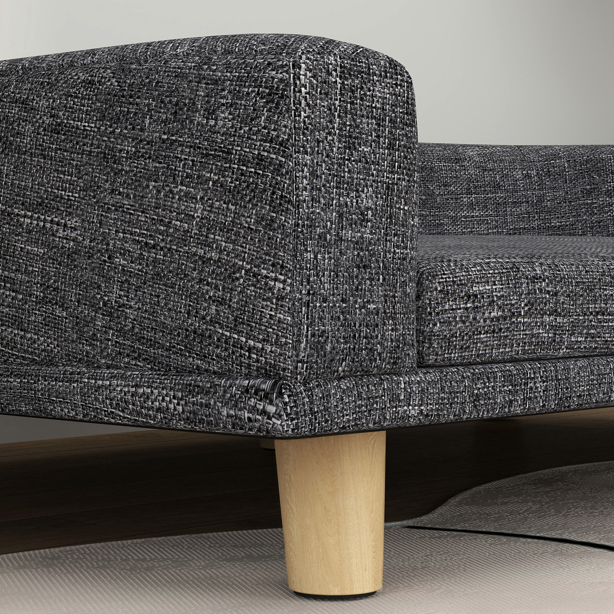 Primer plano de un sofá de tela gris con patas de madera. El sofá está sobre una alfombra estampada y un suelo de madera oscura.