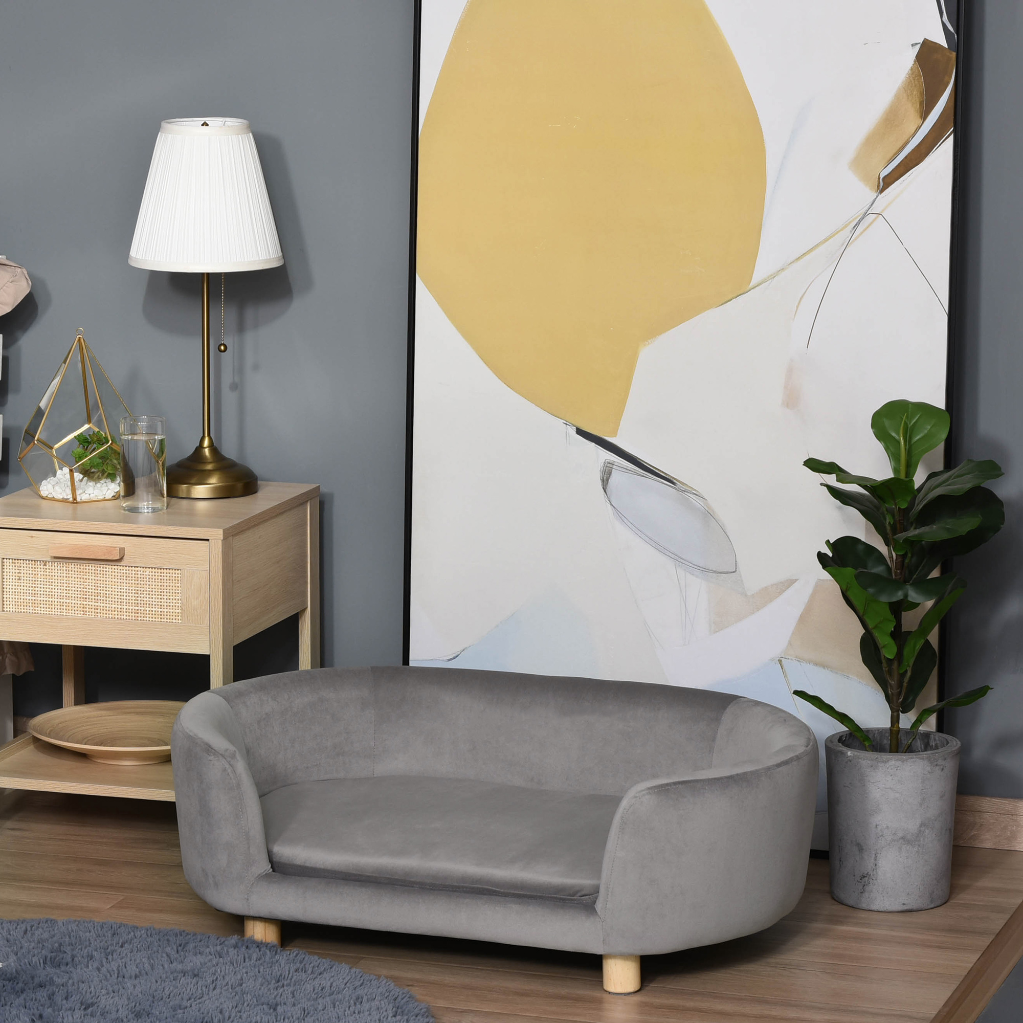Una cama gris para perros en una habitación con pintura, lámpara, planta y muebles de madera.