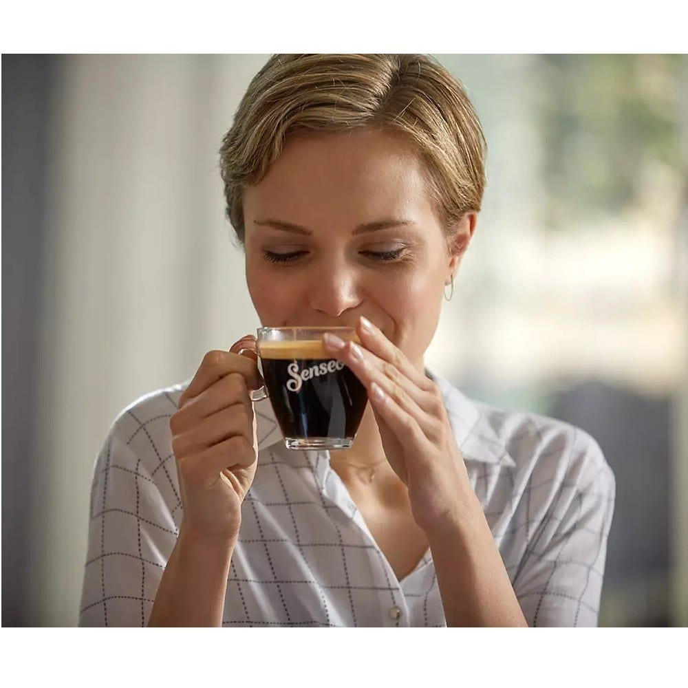 Eine Frau mit kurzen Haaren trinkt Kaffee aus einer Glastasse. Auf der Tasse steht "Senseo". Sie trägt ein weißes Hemd.