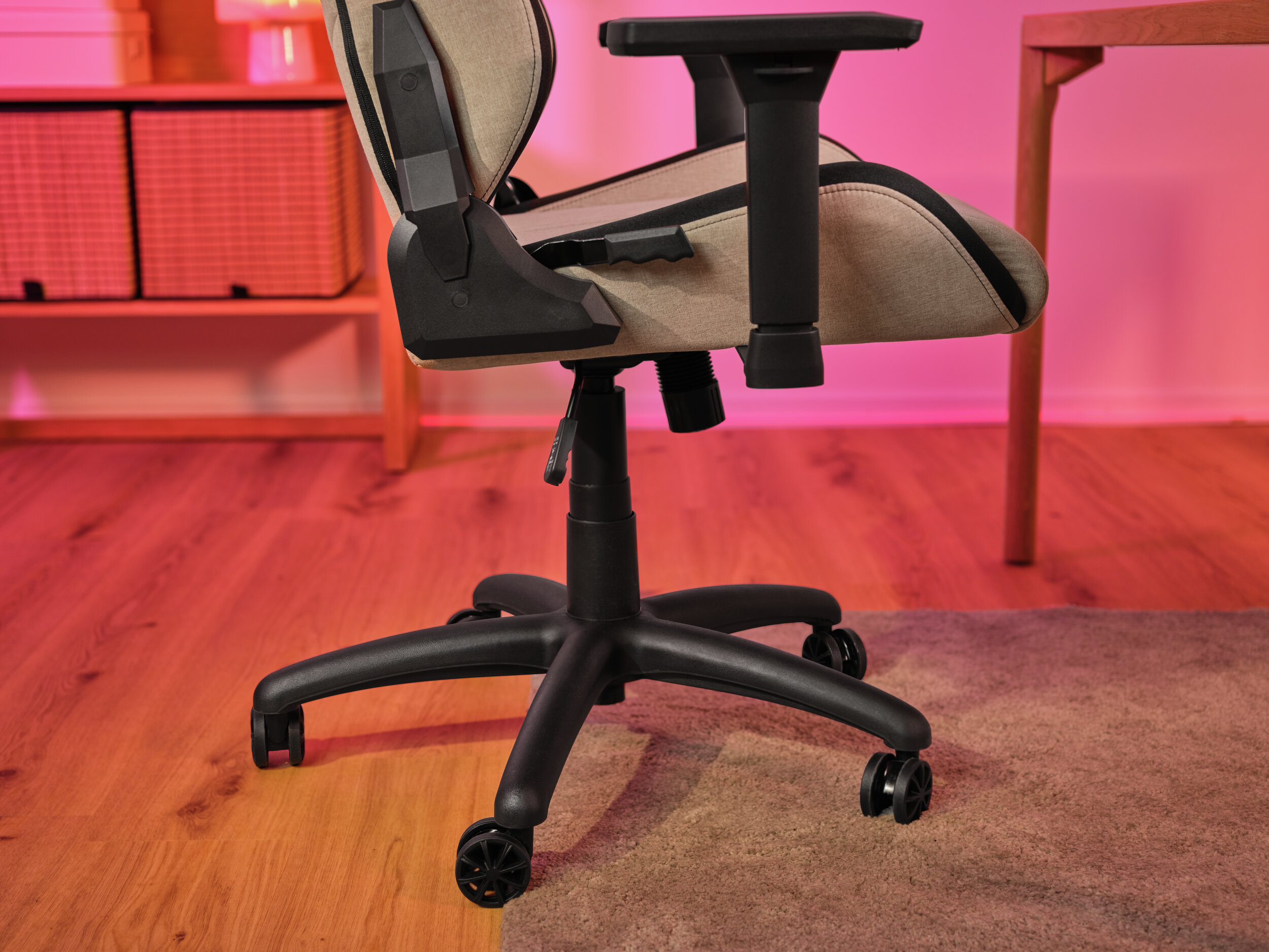 Krzesło do gier z beżowym siedziskiem i czarną ramą stoi na drewnianej podłodze, w pobliżu dywanu. Tło to półka i różowa ściana.