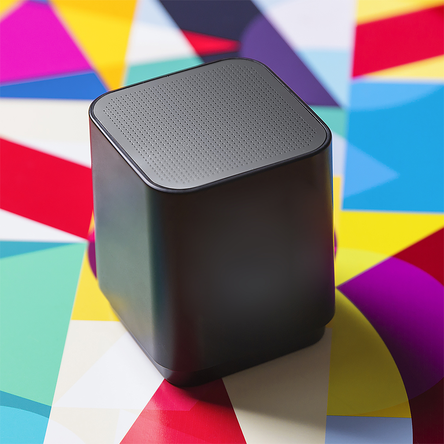 Un altoparlante grigio scuro è posizionato su uno sfondo geometrico colorato di triangoli. L'altoparlante è quadrato e ha una parte superiore perforata.