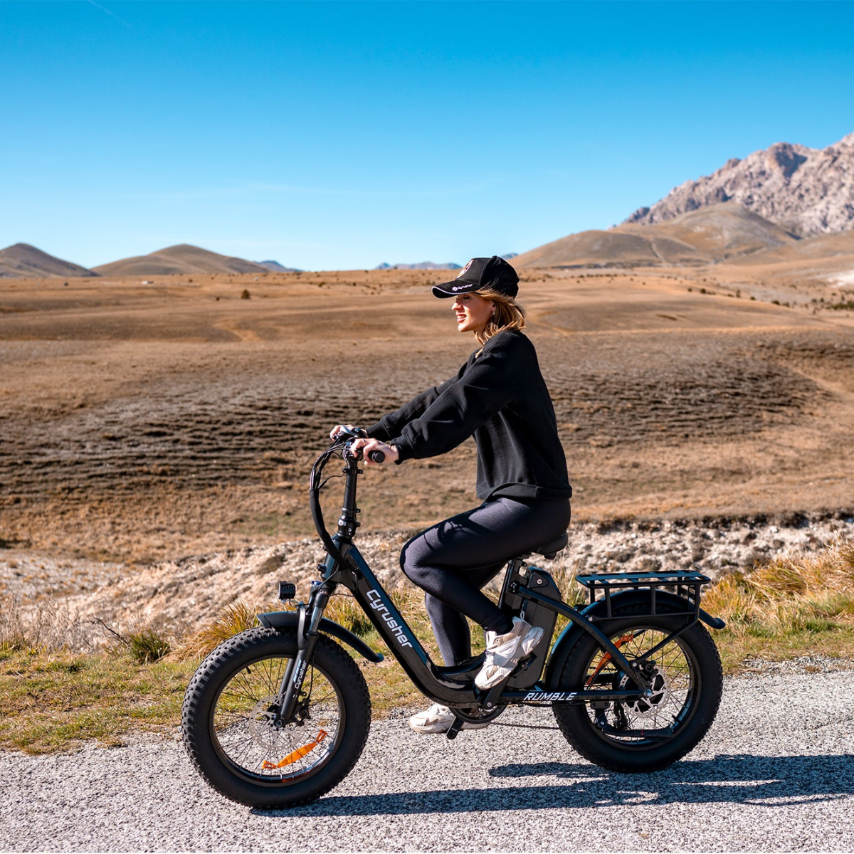 Kobieta jedzie czarnym rowerem elektrycznym po żwirowej drodze, góry w tle pod błękitnym niebem.