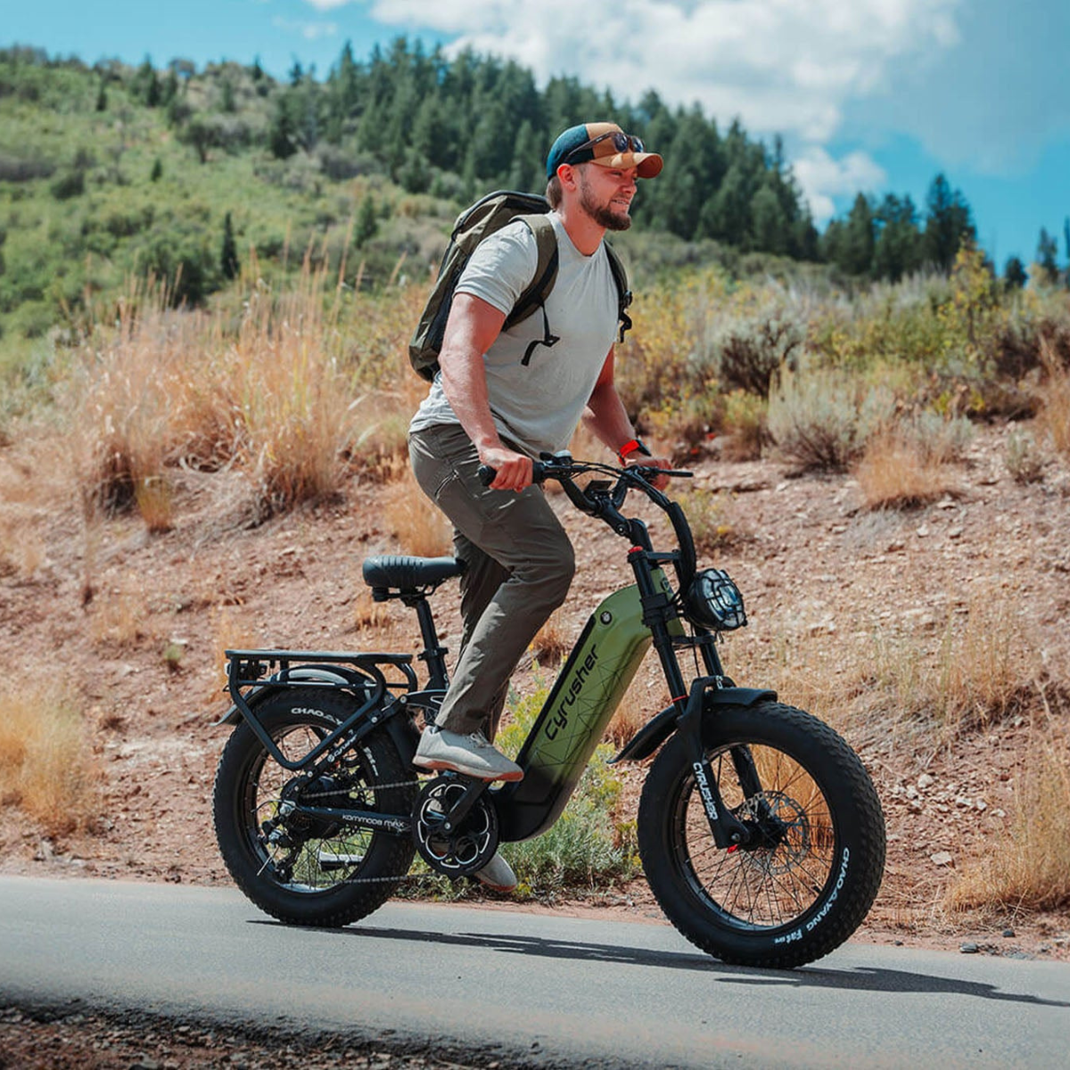 Mężczyzna jedzie na oliwkowozielonym rowerze elektrycznym Cycrusher po utwardzonej drodze. Ma plecak, czapkę i szare spodnie. Tło stanowią wzgórza i drzewa.