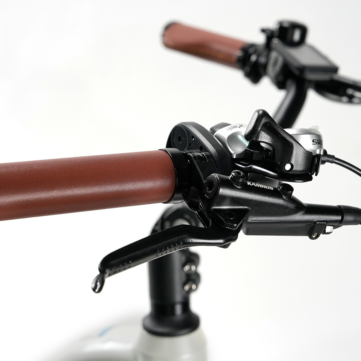 Zbliżenie kierownicy roweru z brązowymi skórzanymi uchwytami, czarnymi dźwigniami i komponentami oraz białym tłem.