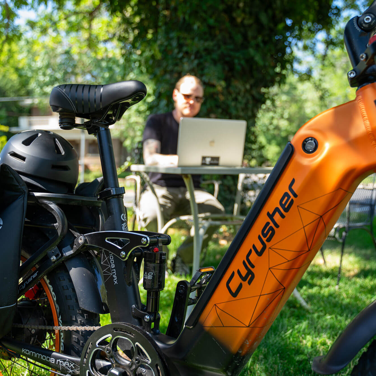 Pomarańczowy rower elektryczny na pierwszym planie. Mężczyzna pracuje na laptopie przy stole w tle, w ogrodzie.