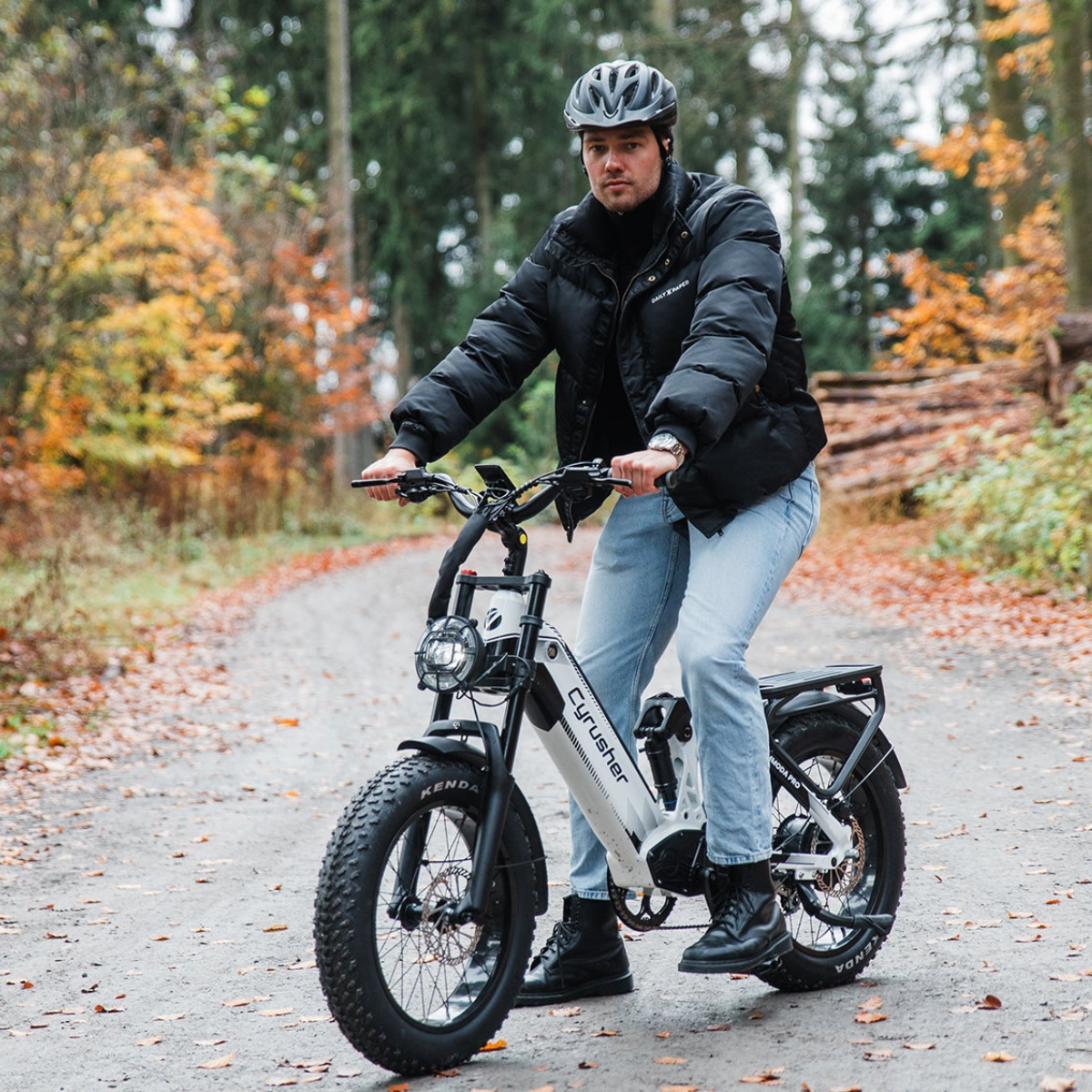 Mężczyzna na białym rowerze elektrycznym w lesie. Ma kask, czarną kurtkę i niebieskie dżinsy. Liście jesienne na ziemi.