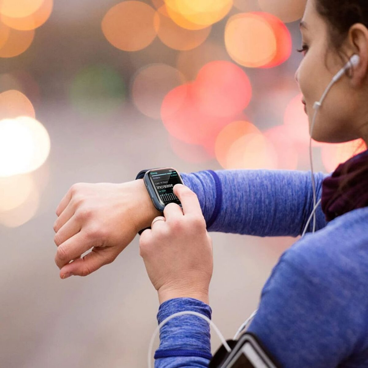 Femme en bleu vérifie smartwatch, écoute de la musique. Arrière-plan flou avec lumières. Focus sur la main et l'écran.