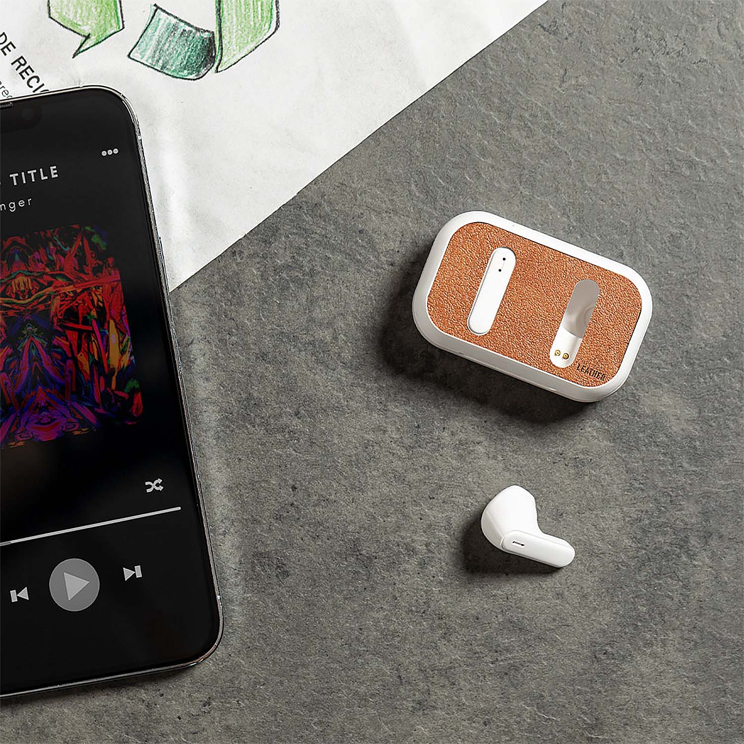 Auricolari, custodia di ricarica e telefono su una superficie grigia. Il telefono mostra l'app musicale. La custodia è in pelle arancione.