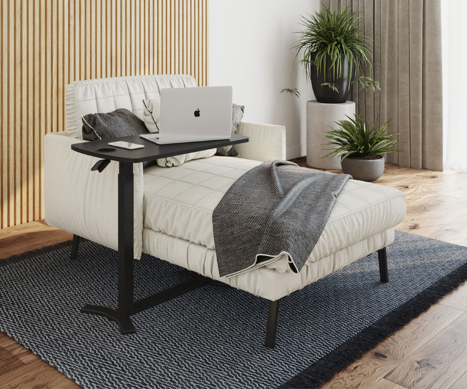 Ein Laptop auf einem schwarzen Tisch steht neben einem weißen Sofa mit einer Decke. Pflanzen in Töpfen sind im Hintergrund. Ein Teppich liegt auf dem Boden.