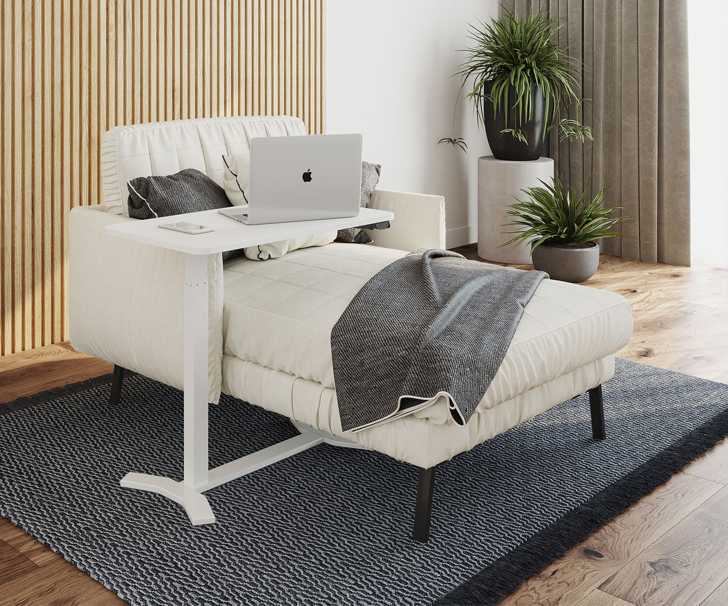 Ein weißer Laptop steht auf einem weißen Tisch über einer cremefarbenen Chaiselongue. Eine graue Decke liegt auf der Liege, Pflanzen im Hintergrund.
