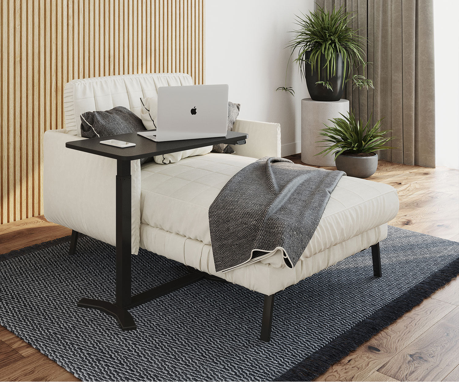 Ein Laptop auf einem schwarzen Tisch neben einer weißen Chaiselongue. Auf der Liege befinden sich eine Decke und Kissen. Im Hintergrund sind Pflanzen.