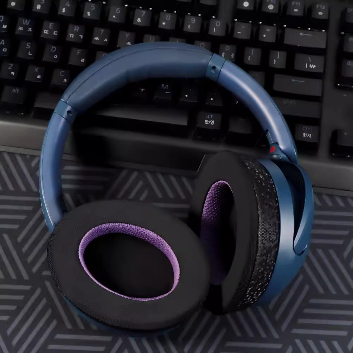 Niebieskie słuchawki na klawiaturze. Nauszniki są czarne z fioletową wyściółką. Tło to czarna klawiatura.