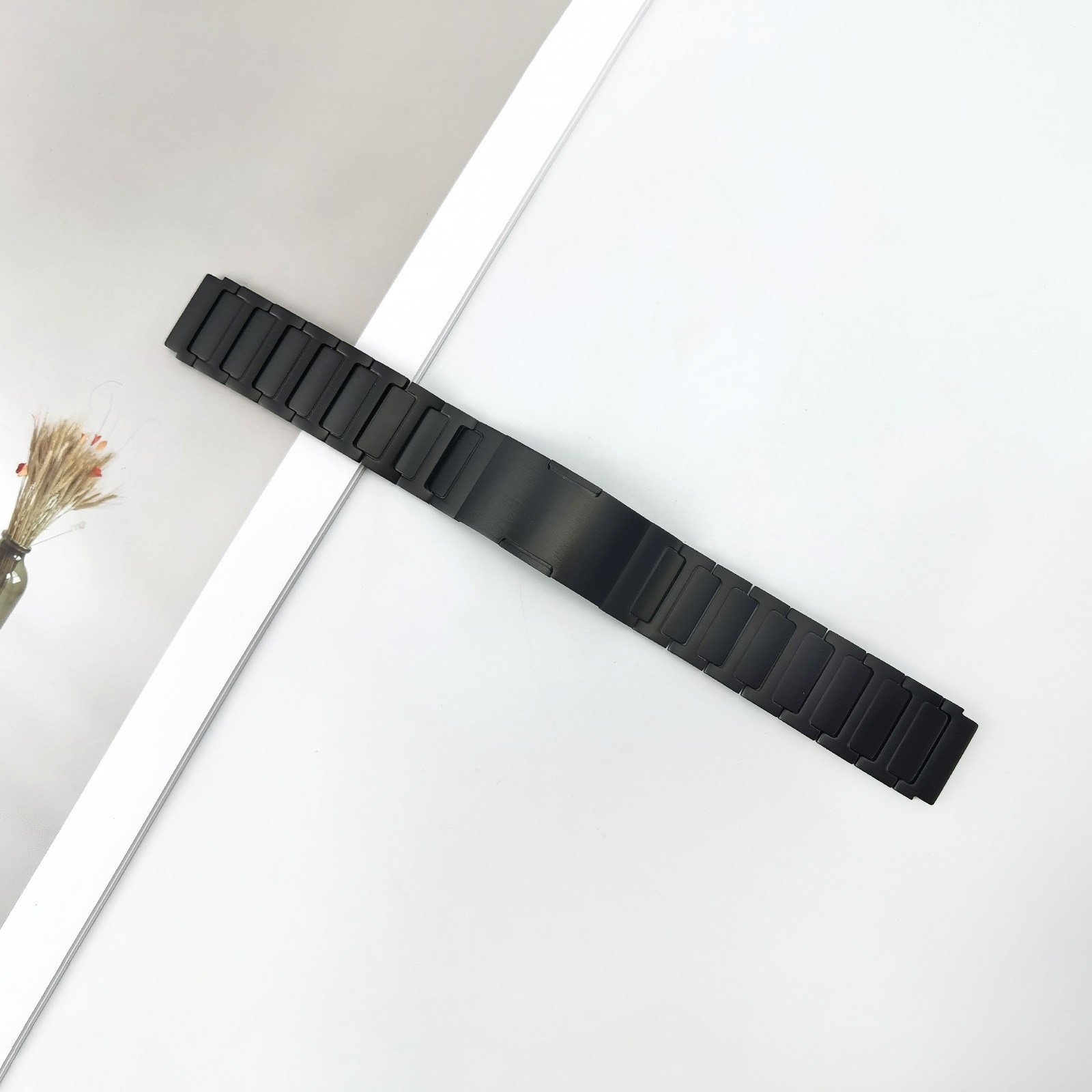 Schwarzes Metall-Uhrenarmband liegt diagonal auf weißem Untergrund. Eine Vase mit Trockenblumen ist im Hintergrund.