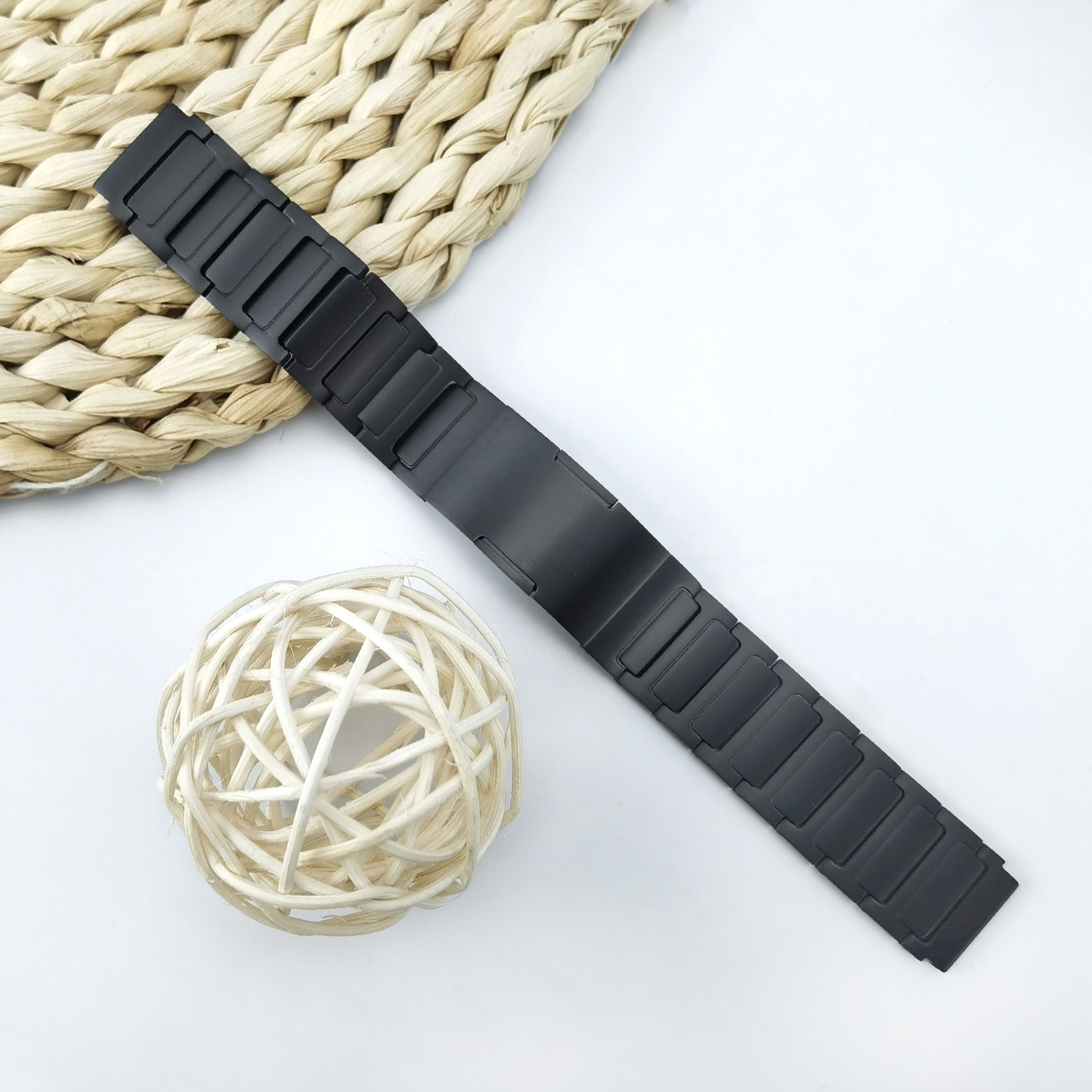 Schwarzes Uhrenarmband auf weißer Oberfläche, mit geflochtenem Stroh und einem Dekoball. Draufsicht.