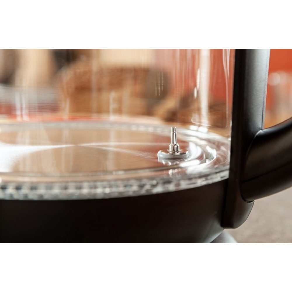 Zbliżenie szklanego czajnika elektrycznego. Mały metalowy element znajduje się na środku. Uchwyt jest czarny, a tło rozmyte.