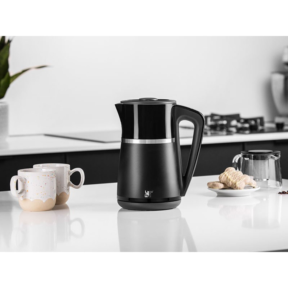 Un bollitore elettrico nero su una superficie bianca. Due tazze e un piatto con biscotti vicini. Cucina sullo sfondo.