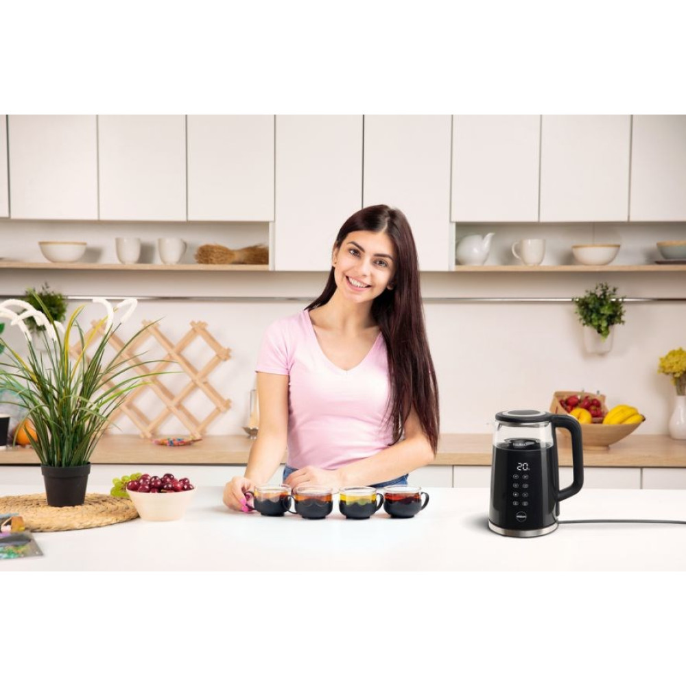 Kobieta w różowej koszuli uśmiecha się, trzyma filiżankę. Czajnik i filiżanki na białym blacie. Rośliny i owoce.