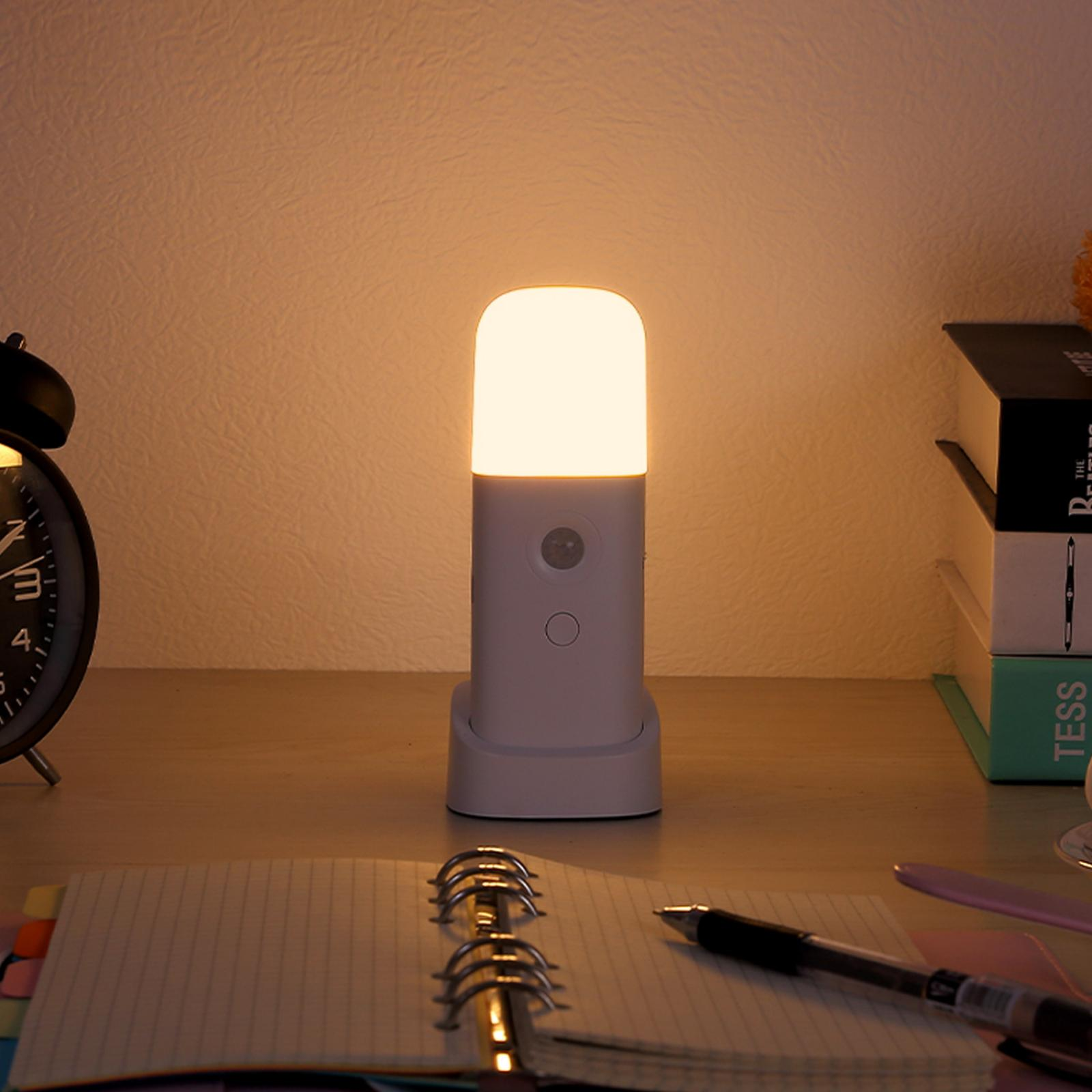 Auf einem Schreibtisch befinden sich eine beleuchtete weiße Lampe, eine Uhr, ein Notizbuch und Bücher. Die Lampe steht im Mittelpunkt.