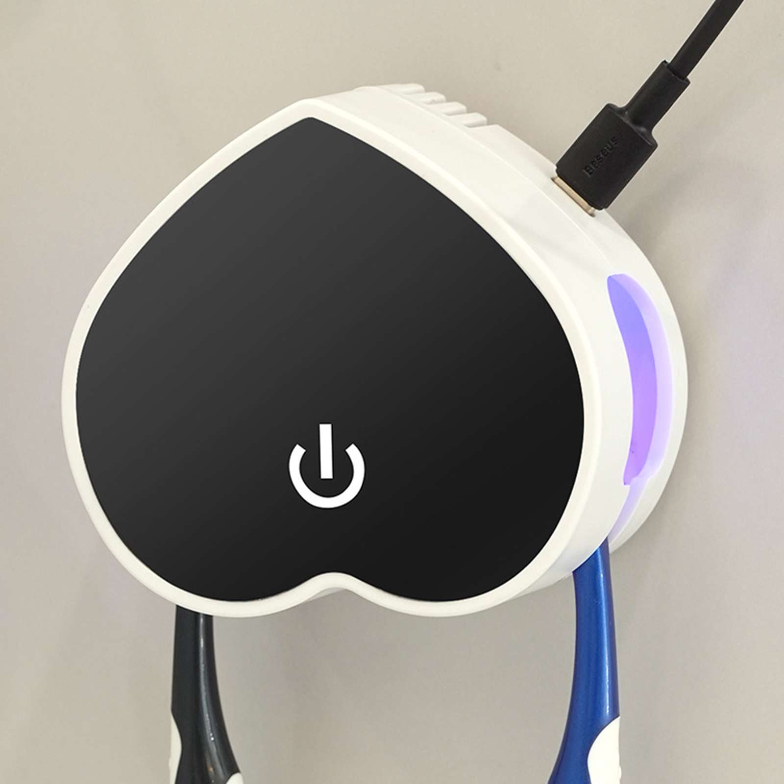Weißes herzförmiges Gerät mit schwarzem Mittelteil und Ein-/Aus-Taste. Schwarzes USB-Kabel eingesteckt. Zwei Zahnbürsten unten.