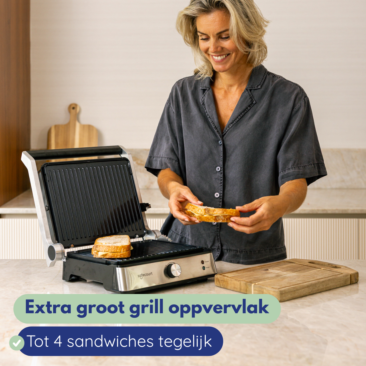 Woman smiles, holding a sandwich near a grill with a sandwich inside. A wooden cutting board is on the counter.