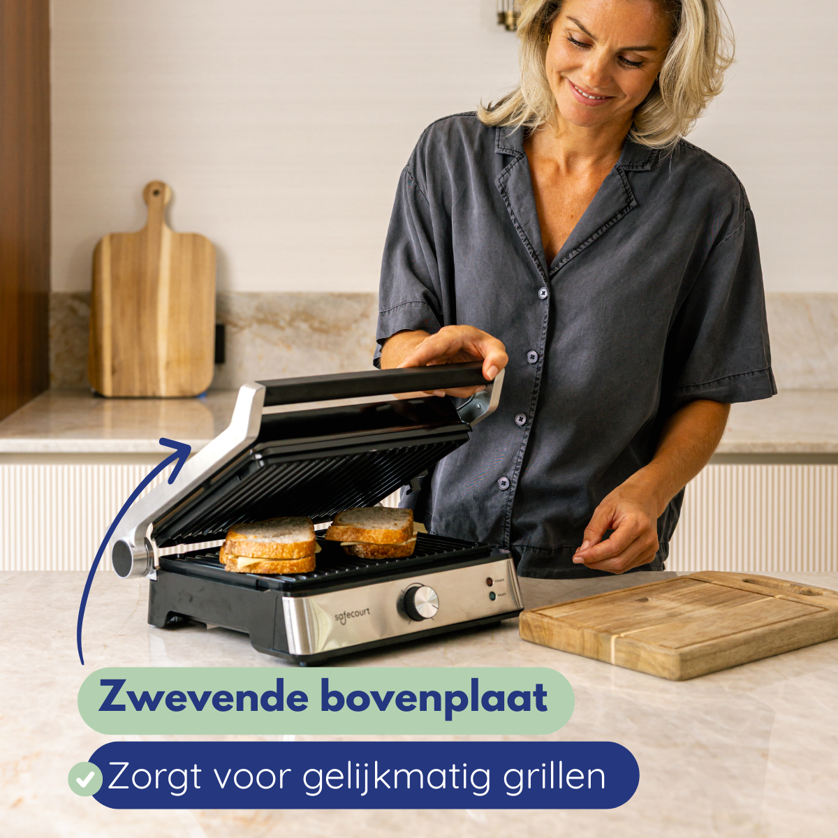 A woman using a sandwich grill in a kitchen. Two sandwiches are inside. A cutting board is nearby. The woman smiles.