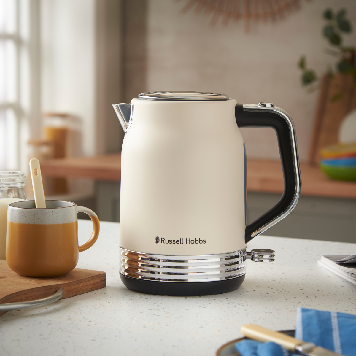 Ein cremefarbener Wasserkocher von Russell Hobbs steht neben einer Tasse auf einem Holzbrett. Im Hintergrund sind Küchenzeile und Utensilien zu sehen.