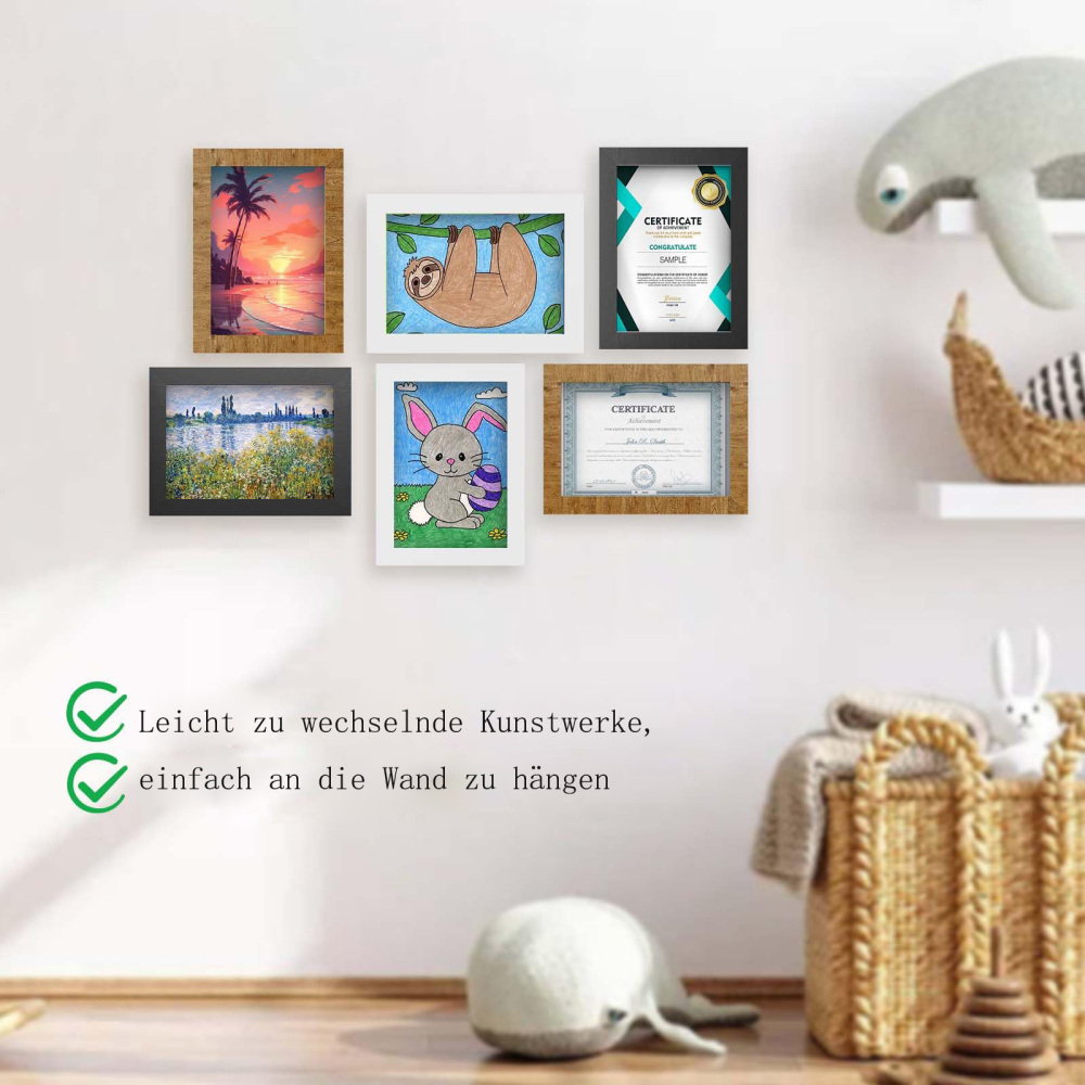 Wand mit gerahmter Kunst: Sonnenuntergang, Faultier, Zertifikat, Hase mit Ei und Landschaft. Ein Plüschwal und Korb mit Spielzeug sind davor.