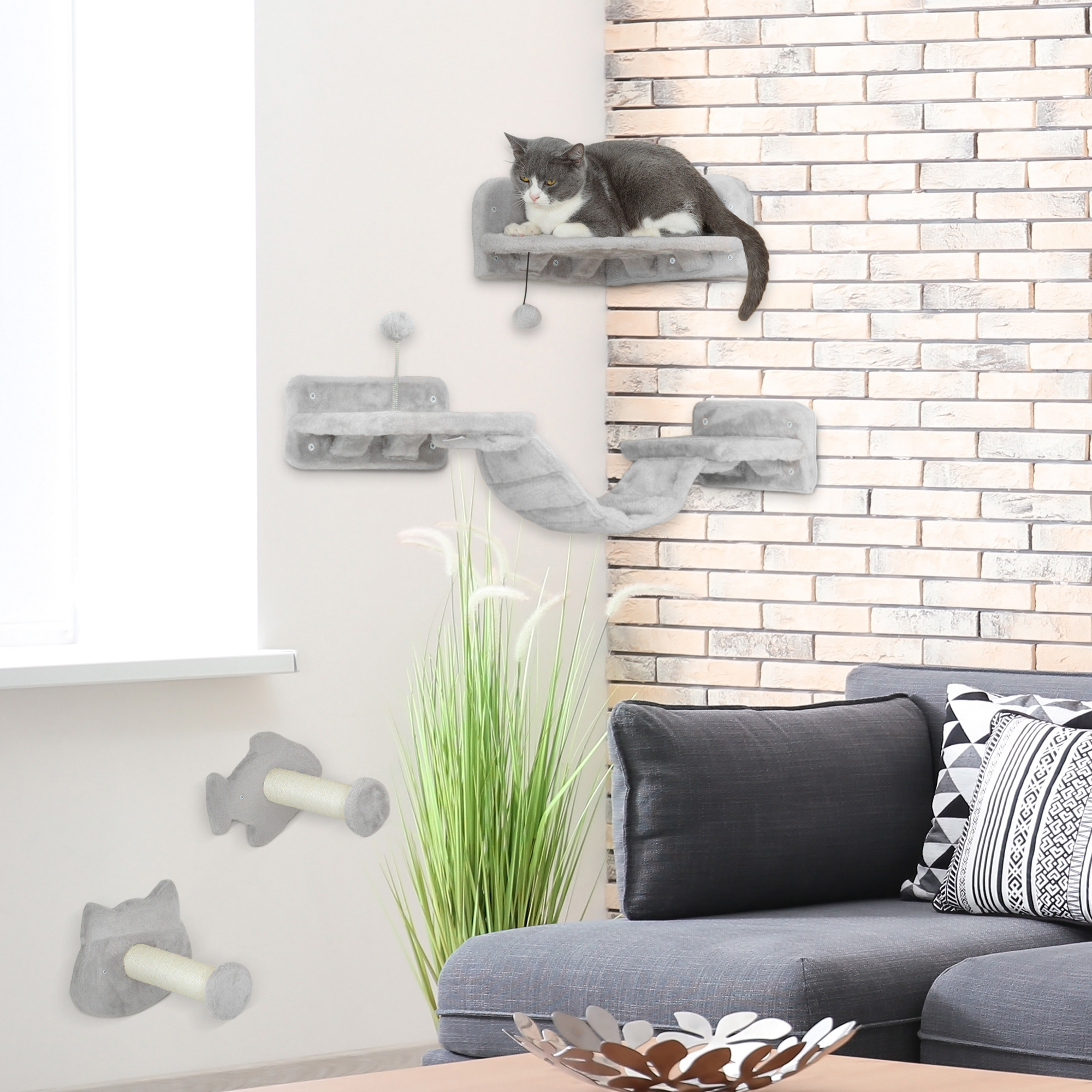 Un gato gris y blanco descansa sobre una cama para gatos montada en la pared. La escena incluye muebles para gatos, un sofá, pared de ladrillos y plantas.