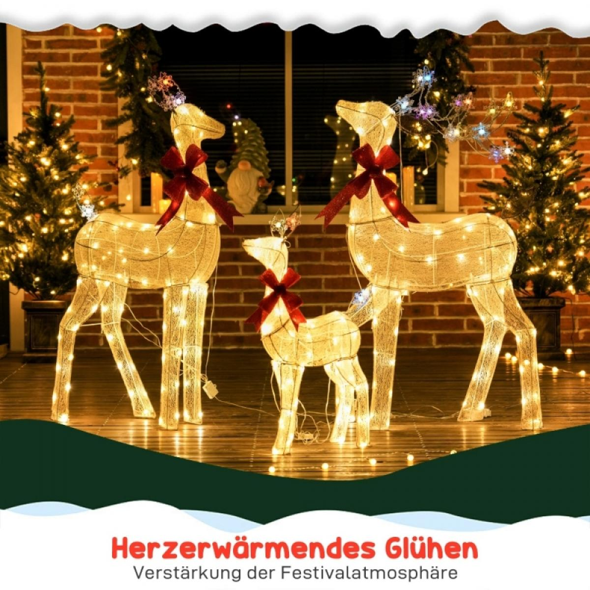 Beleuchtete Rentierdekorationen mit roten Schleifen stehen vor einer Backsteinmauer und Weihnachtsbäumen. Die Szene ist festlich und warm.