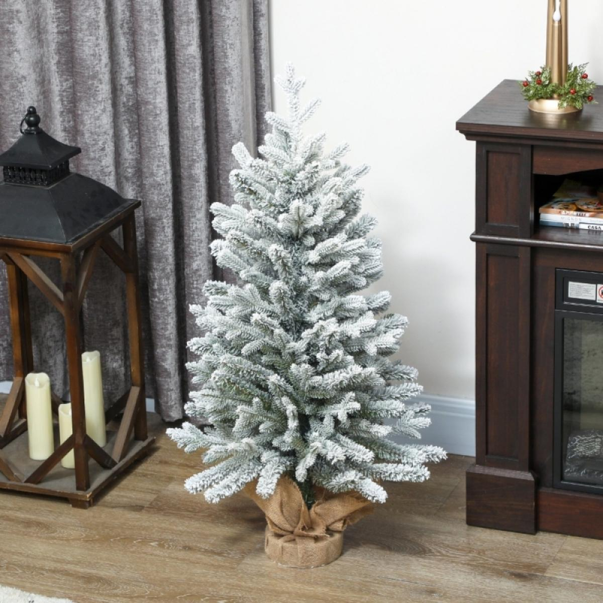 Ein kleiner Weihnachtsbaum mit schneeglasiertem Frost steht auf einem Holzboden. Eine Laterne und ein dunkler Schrank sind im Hintergrund.