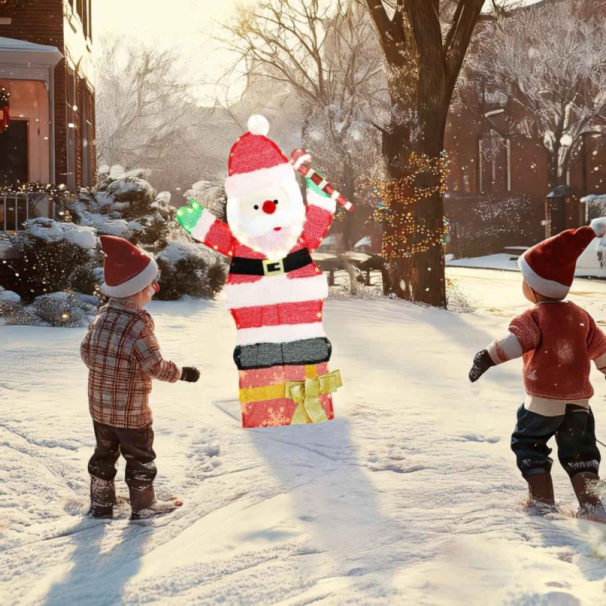 Zwei Kinder spielen im Schnee neben einer Weihnachtsmann-Dekoration. Im Hintergrund sind Häuser und Bäume zu sehen, eine sonnige Winterszene.