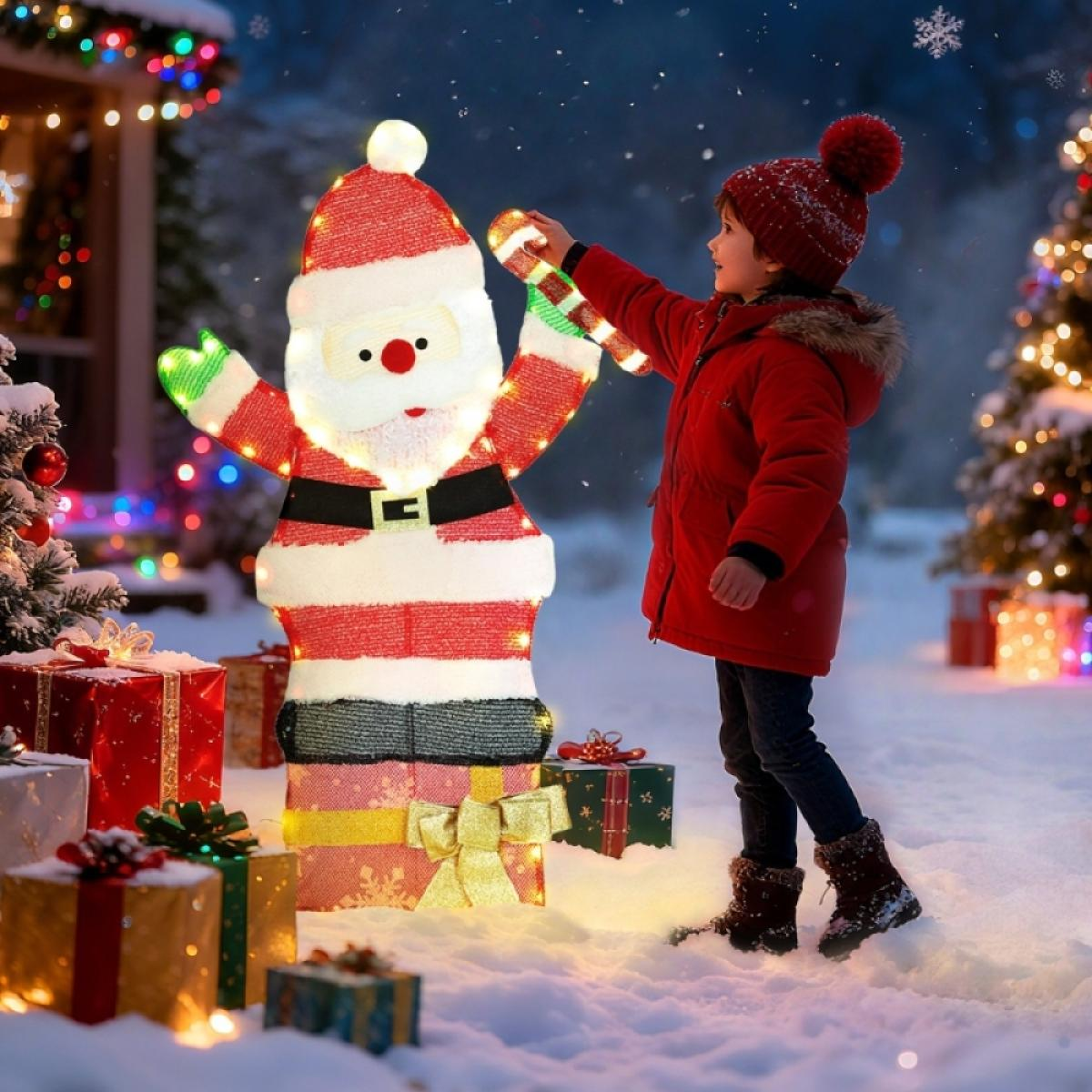 Ein Kind in roter Jacke und Mütze schmückt eine beleuchtete Weihnachtsmannfigur. Geschenke und ein Weihnachtsbaum sind im schneebedeckten Hintergrund.