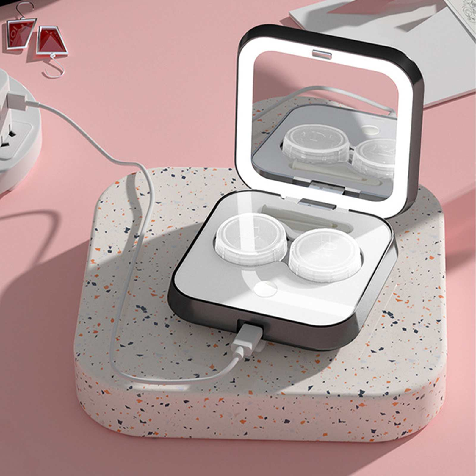Kontaktlinsenbehälter mit Spiegel auf gesprenkelter Oberfläche. Rosa Hintergrund, Netzkabel und Stecker sichtbar.