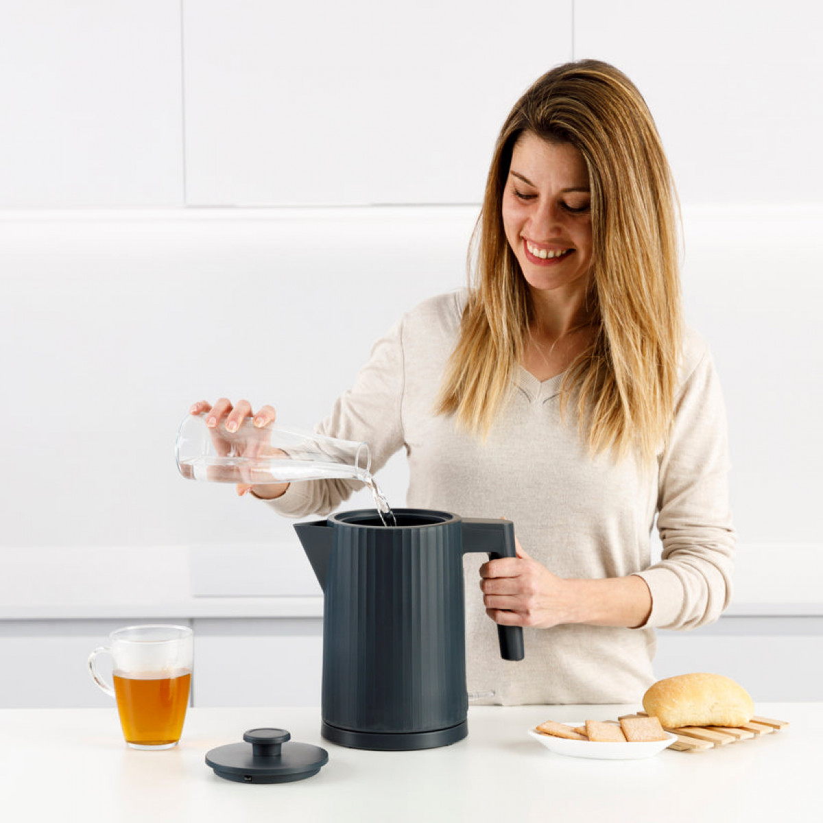 Kobieta wlewa wodę ze szklanki do ciemnoszarego czajnika elektrycznego. Na stole filiżanka herbaty, chleb i krakersy.