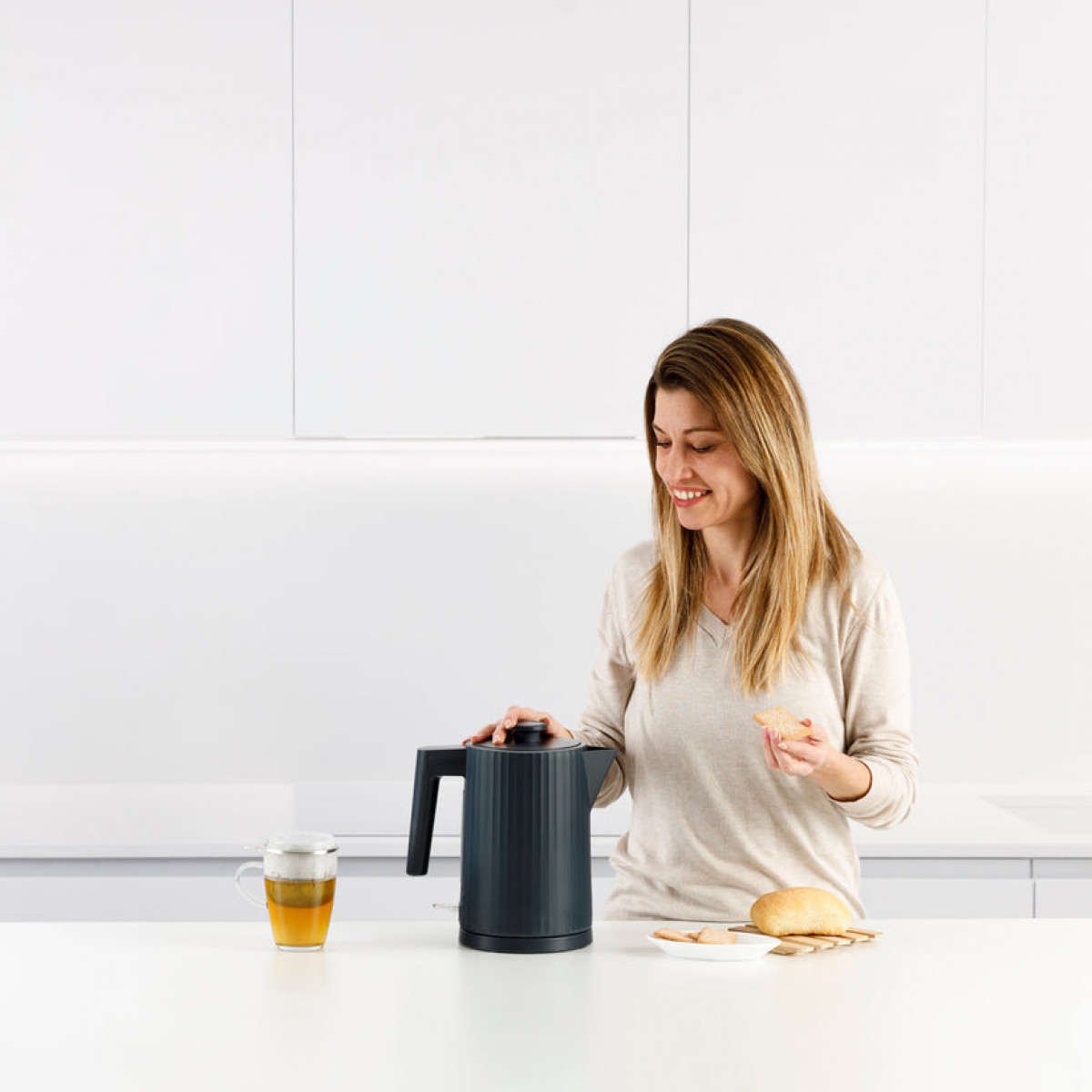 Kobieta uśmiecha się, trzymając ciastko, obok czajnika, filiżanki herbaty i chleba na białym blacie. Tło jest białe.