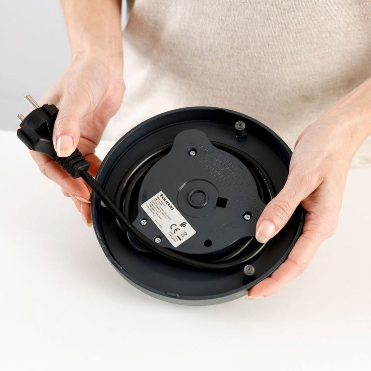 Osoba trzyma podstawę czarnego czajnika, pokazując kabel zasilający i wtyczkę. Podstawa jest ciemnoszara.