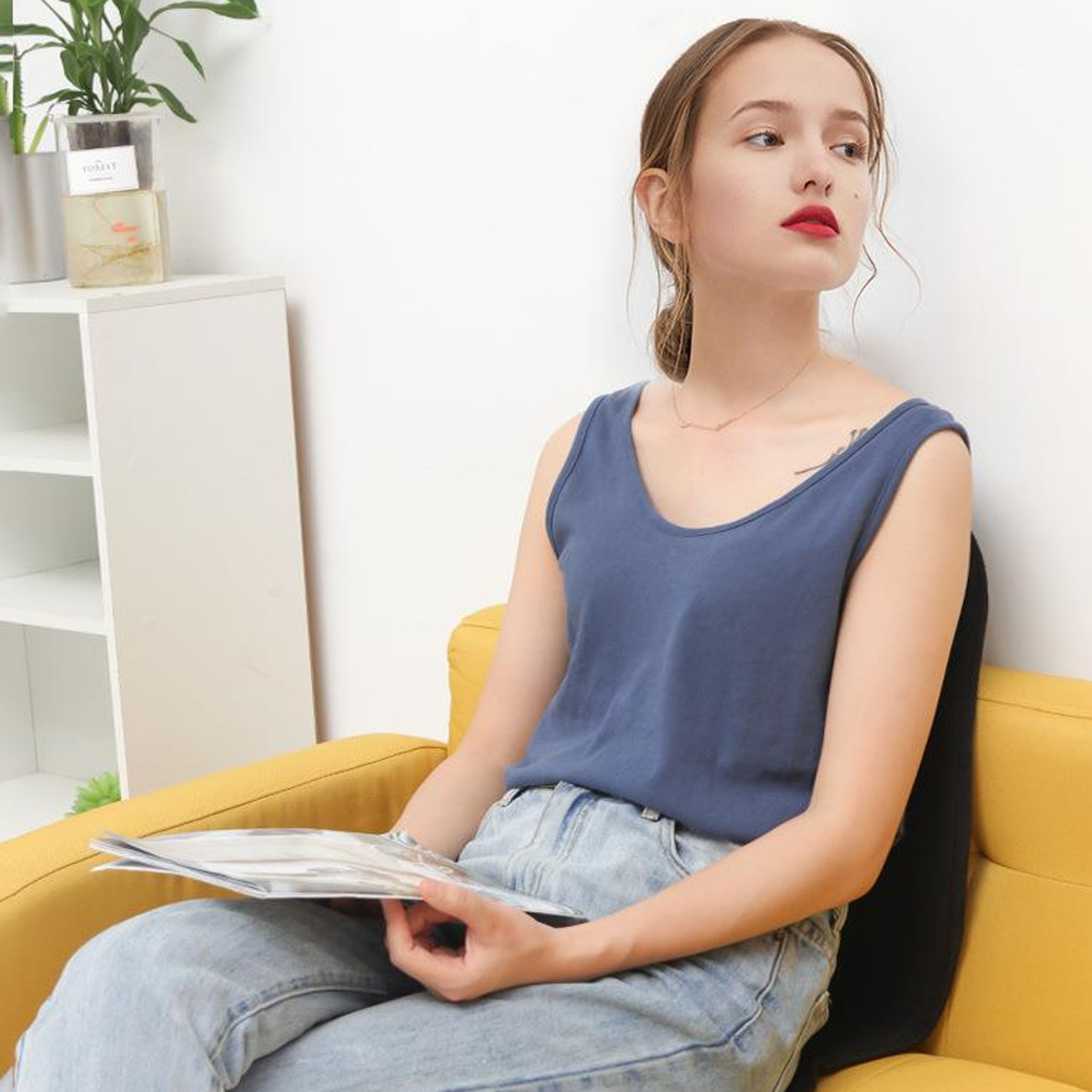 Eine Frau in einem blauen Tanktop und Jeans sitzt auf einem gelben Sofa und hält eine Zeitschrift. Sie schaut nach rechts. Links ist eine Pflanze.