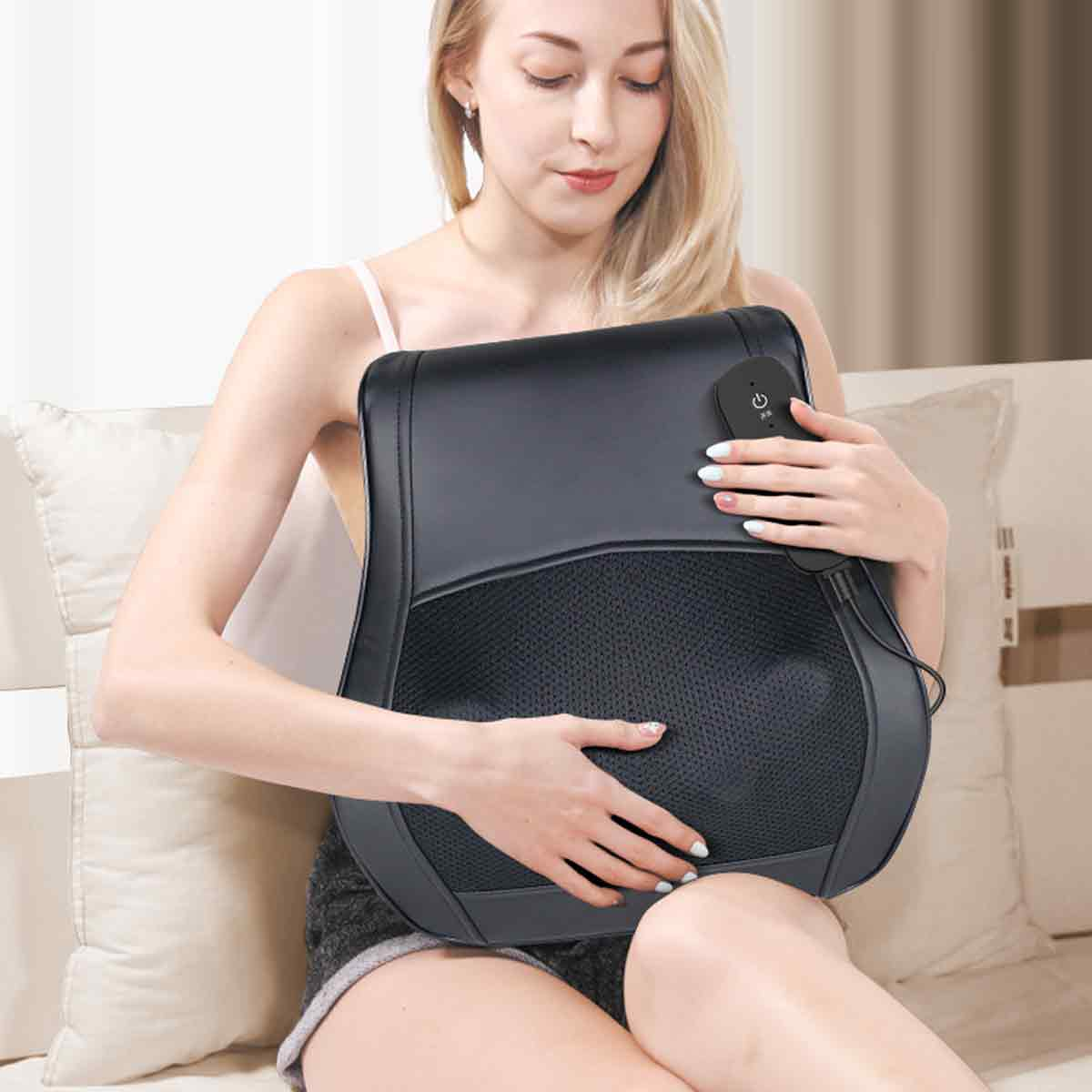 Eine Frau mit blonden Haaren hält ein schwarzes Massagekissen. Das Kissen liegt an ihrem Bauch. Sie sitzt auf einem Bett mit weißen Kissen.