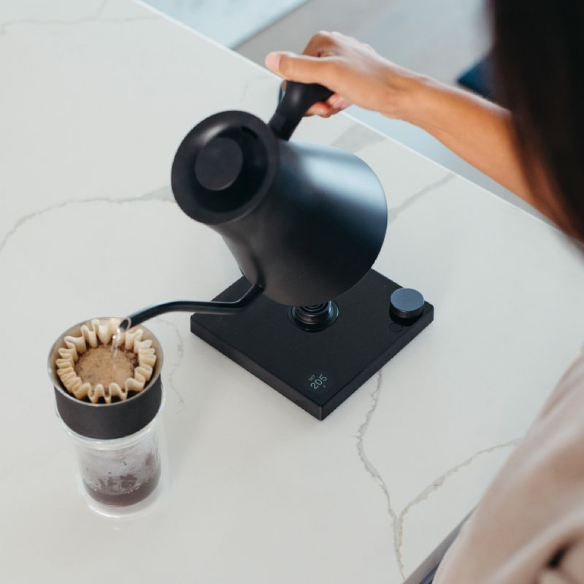 Osoba wlewa wodę z czarnego czajnika do filtra do kawy. Scena rozgrywa się na białym blacie z ekspresem do kawy.