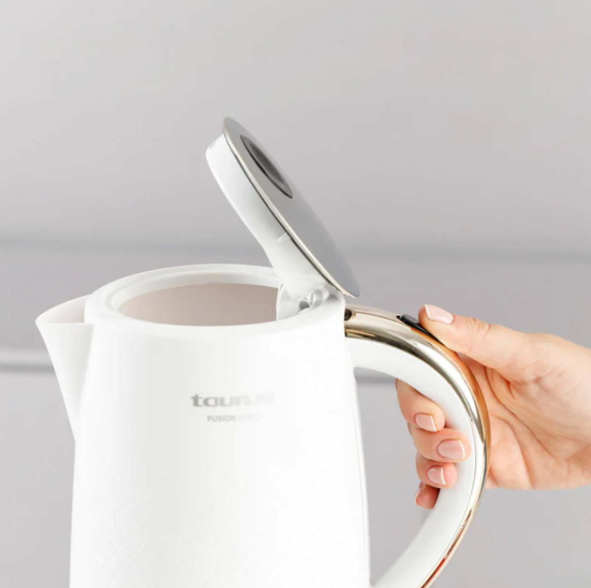 Biały czajnik elektryczny z otwartą pokrywką. Dłoń trzyma uchwyt. Czajnik ma złoty i biały uchwyt oraz srebrną pokrywę.