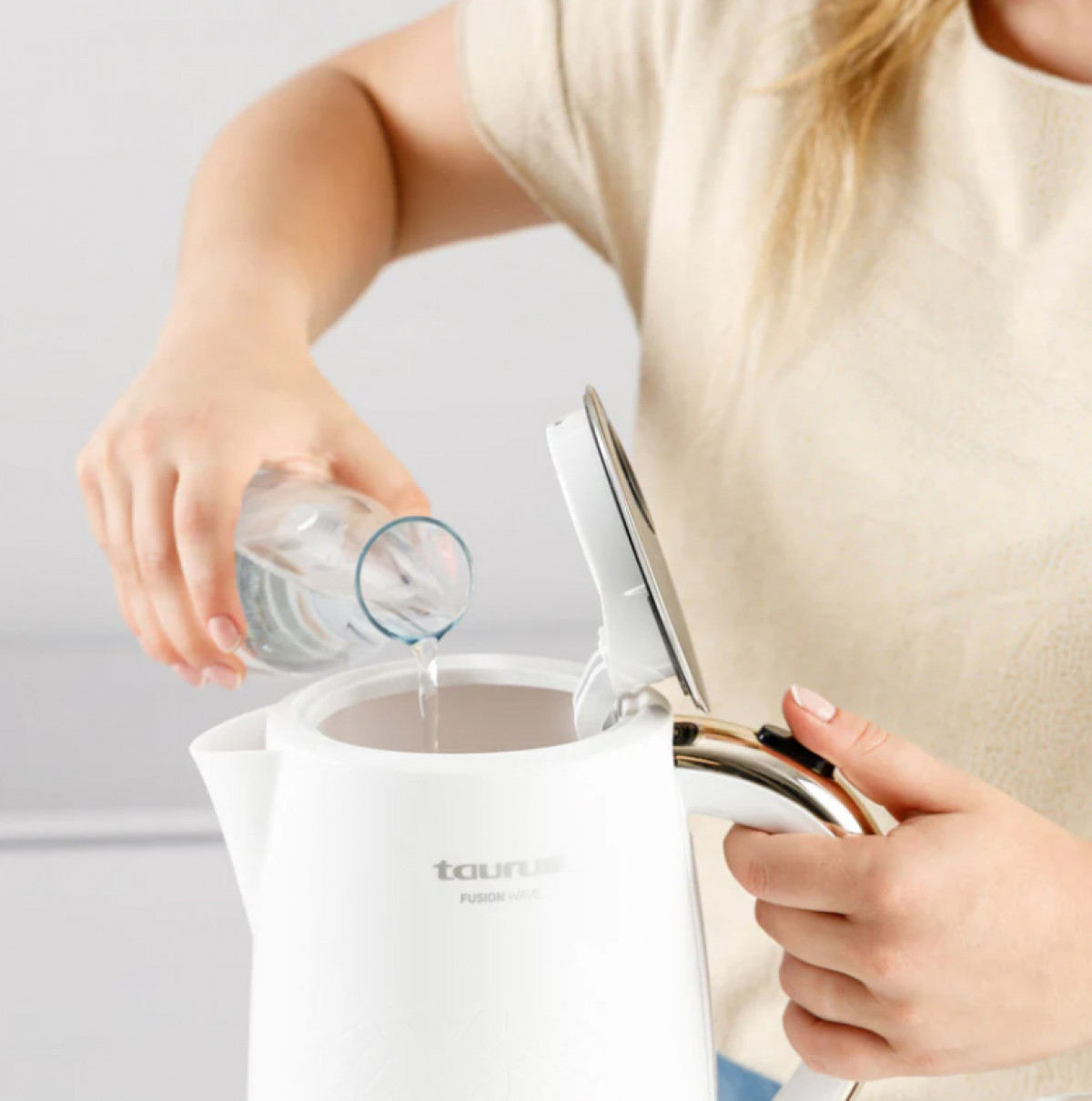 Kobieta wlewa wodę z butelki do białego czajnika. Pokrywa czajnika jest otwarta. Kobieta ma na sobie jasną koszulę.