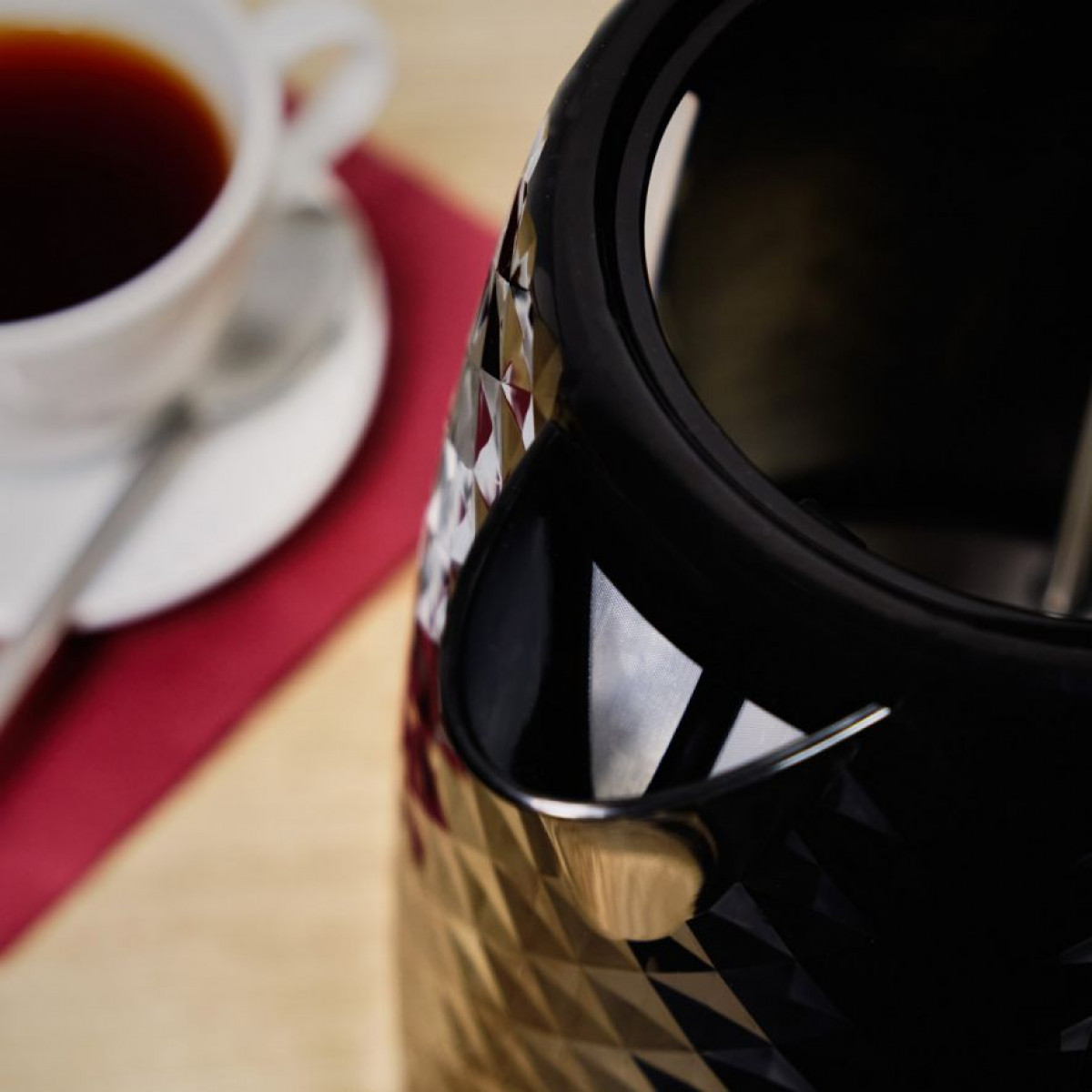 Zbliżenie czarnego czajnika elektrycznego z białą filiżanką i spodkiem do kawy na czerwonej serwetce.