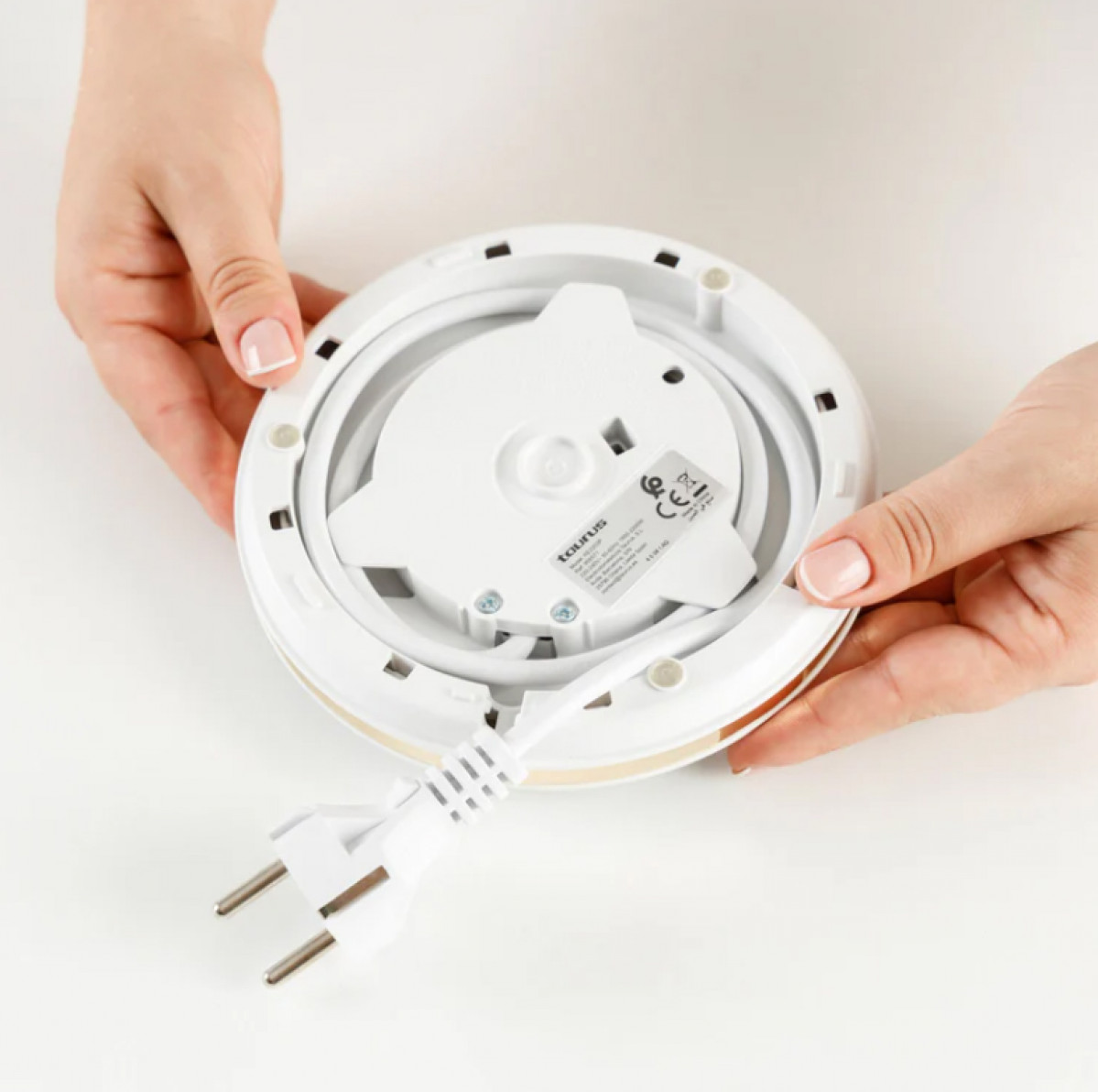 Ręce trzymają biały, okrągły czajnik elektryczny. Biały przewód zasilający i wtyczka wychodzą z czajnika. Tło jest białe.