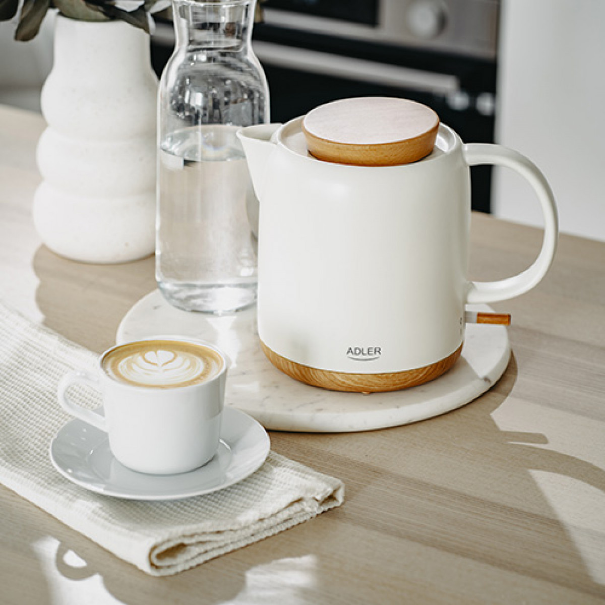 Biały czajnik elektryczny z drewnianymi akcentami, filiżanka kawy, wazon i szklana butelka na drewnianej powierzchni.