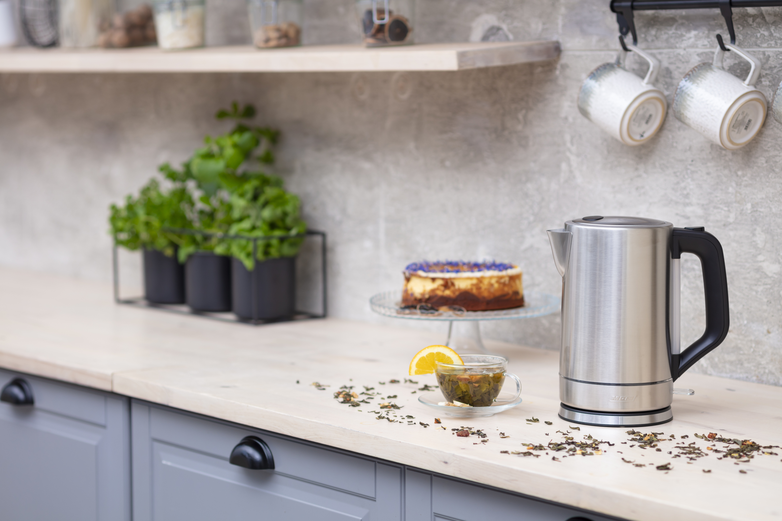 Una scena di cucina con un bollitore, tè, torta e tazze. Piante verdi e una mensola di legno sullo sfondo.