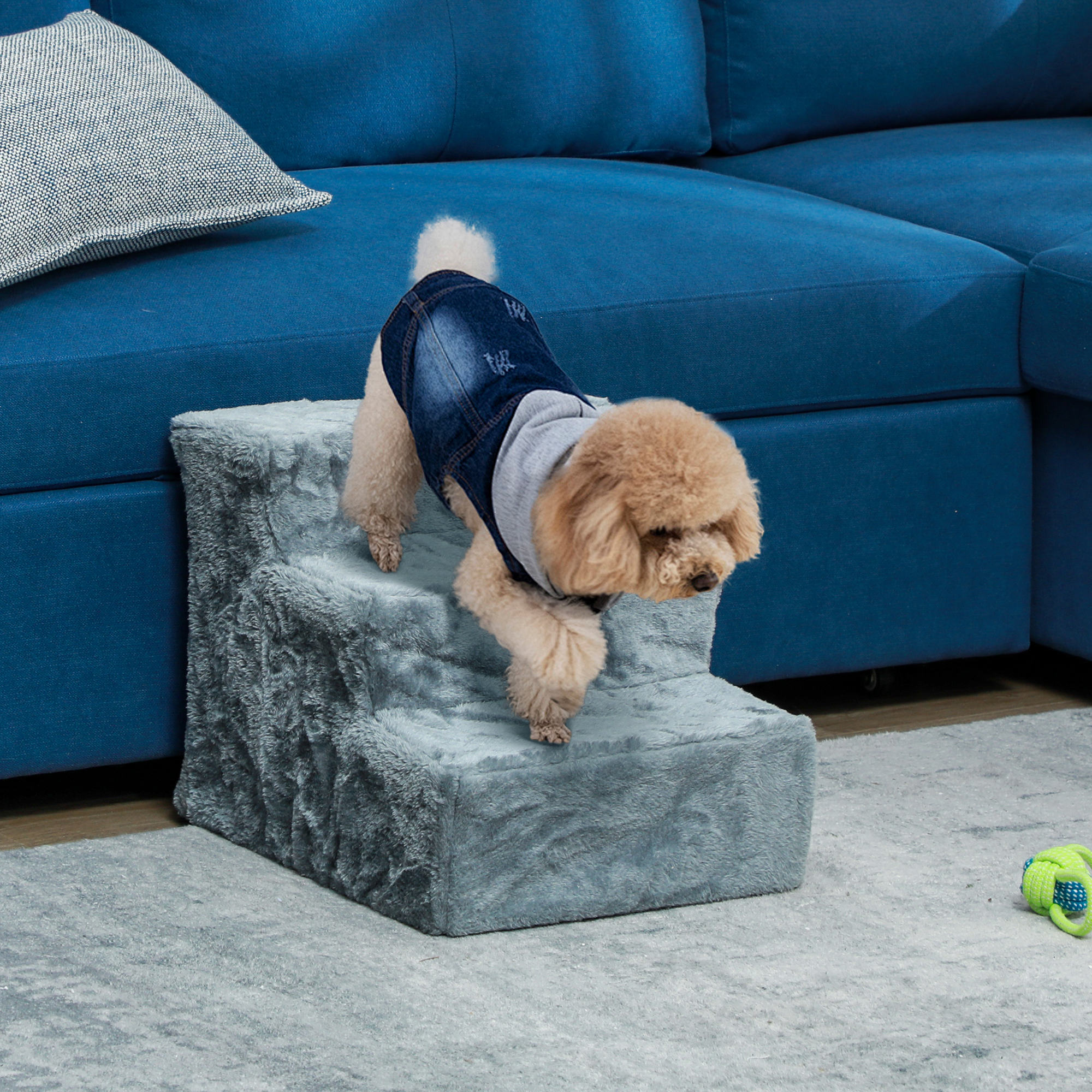 Un perro pequeño con una chaqueta vaquera desciende por unas escaleras grises para mascotas. Un sofá azul está al fondo, un juguete en el suelo.