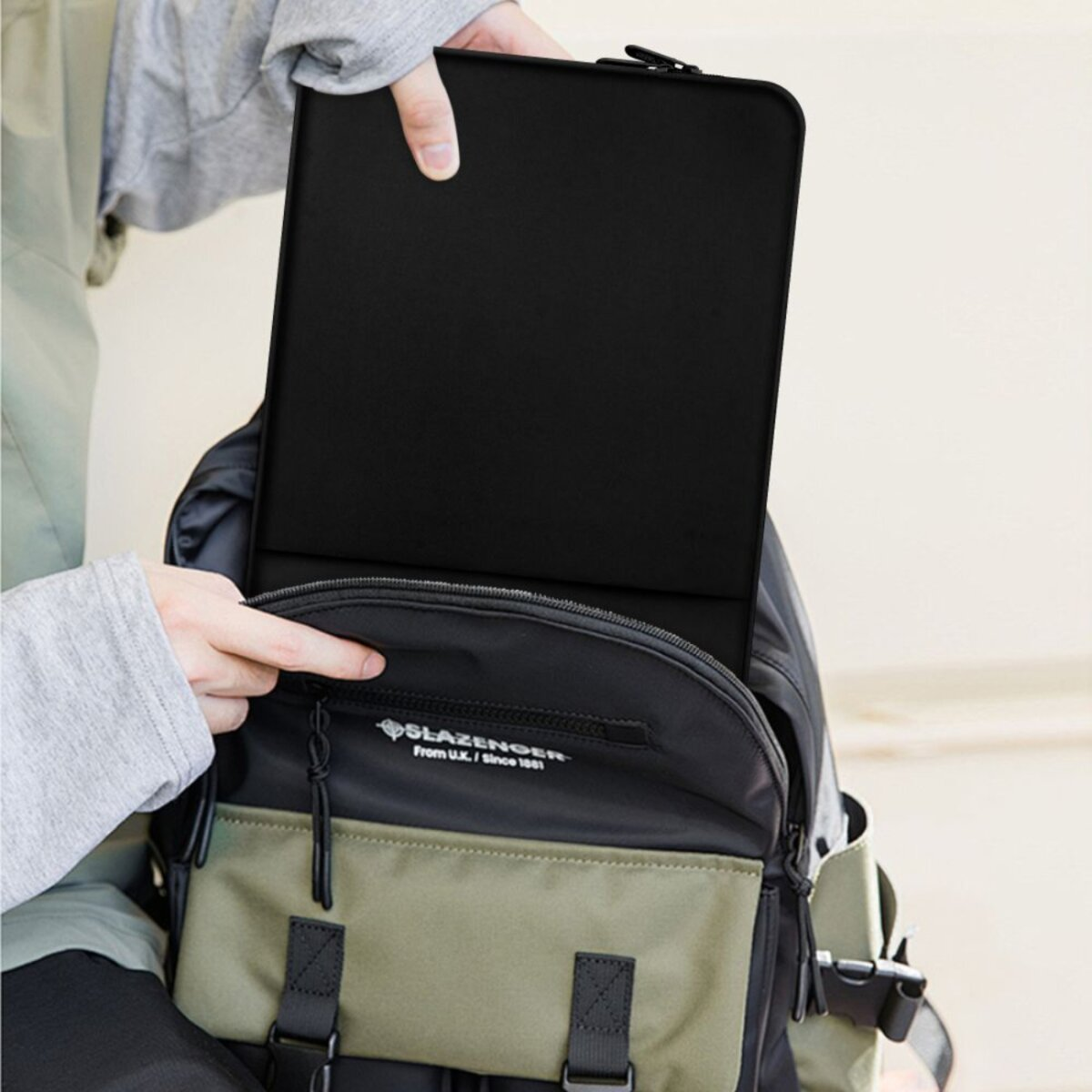 Een persoon stopt een zwarte tablet in een rugzak. De rugzak is zwart en groen, met een Slazenger-logo.
